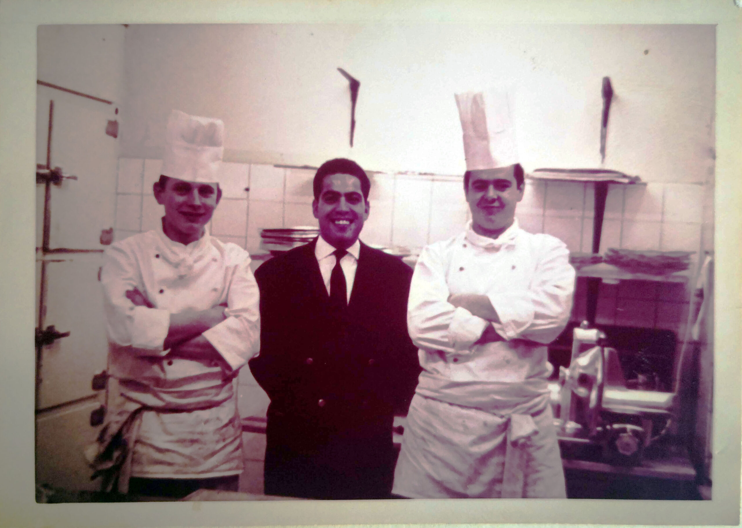 Vintage photo of Gert and friends in the kitchen, two wearing chef hats and one in a suit, showcasing culinary heritage