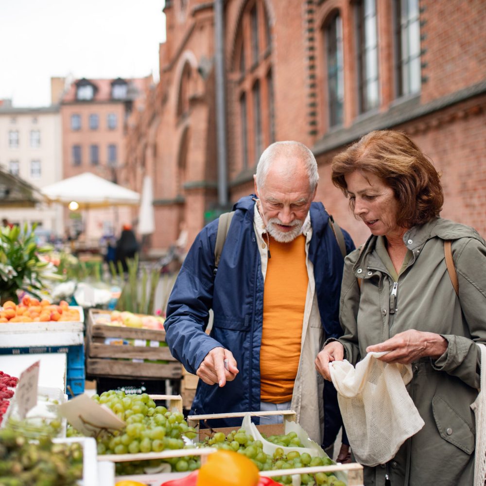 A happy senior couple tourists buying fruit outdoors on market in town.